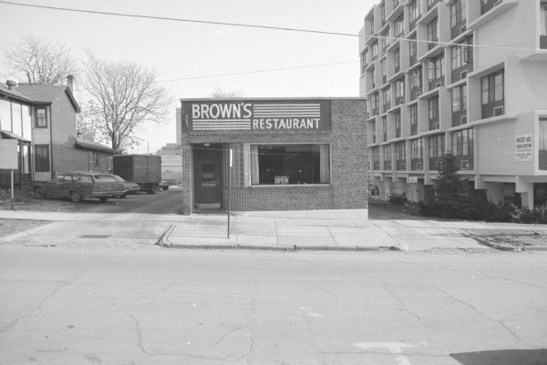 View across West Gilman Street toward the Floyd Brown Building, which contained Brown's Restaurant. A sign in the window reads: "Yes We're Open." A sign on the door lists their business hours. To the right of Brown's Restaurant is Master Hall, an apartment complex. Cars, and a van for Stop 'n Shop Grocery are parked in the lot near buildings on the left, next to a store called Lady Cybele's Cauldron.
