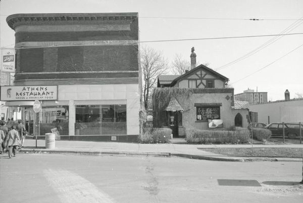 View across West Gilman Street, on the corner of State Street and Broom Street, towards two commercial buildings. The one on the left is the Stephensen and Studemann Commercial Building, which contains Athens Restaurant. The building on the right is the James Murtah House. The building has vines growing on the facade over the entrance, and contains Lady Cybele's Cauldron. Decals in the window read: "Lady Cybele's Cauldron: Occult Supplies & Services, Books, Jewelry." To the right of Lady Cybele's is the side of Brown's Restaurant. A group of people are walking across the street towards Athens Restaurant. 