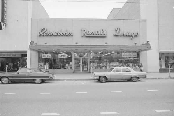 View across East Main Street towards the Rennebohm Drug Store. Pedestrians are walking on the sidewalk, and two cars are parked along the curb. Signs in the show windows are advertising a "Soda Grill," "Cafeteria," "Fine Wines & Liquors," and various other goods.