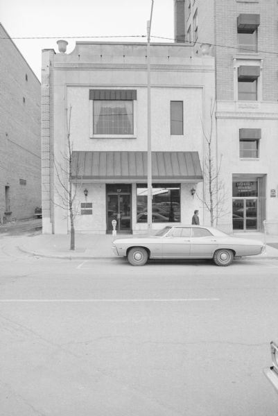 View across Monona Avenue (now Martin Luther King Jr Boulevard) towards the Stark Company Realtors. The roofline has decorative urns at the corners. An automobile is parked in front of the building, and a man is walking on the sidewalk. To the right is an insurance building at 119 Monona Avenue. 