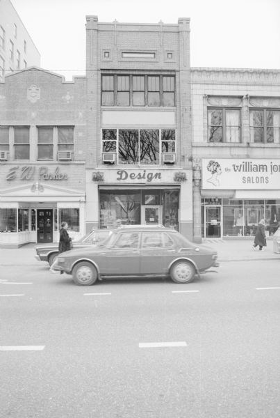 View across West Main Street towards the three-story J.J. Coppernell Business Block, which includes the Design Art Gallery Boutique. The facade has decorative brickwork, and the ground floor has show windows flanking the entrance. On the left is E.W. Parker Jewelry, and on the right is The William Jon Salons. A woman is walking on the street near a parked car and another car being driven down the street.