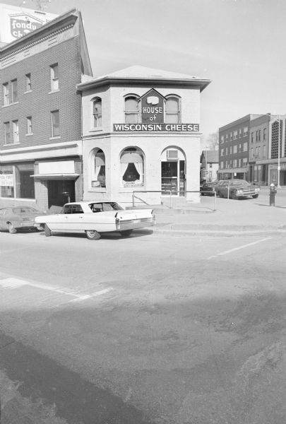 View across North Hamilton Street, on the corner of East Mifflin Street, towards the Draper Brothers Block. A large neon sign above the door reads: "House of Wisconsin Cheese." Various cheese products are on display in the window, and more can be seen through the door. A large billboard is partially visible above the building, and advertises Fondue Cheese. The building adjacent to House of Wisconsin Cheese has a sign in the ground floor window indicating it is for rent. Cars are parked along the curb.