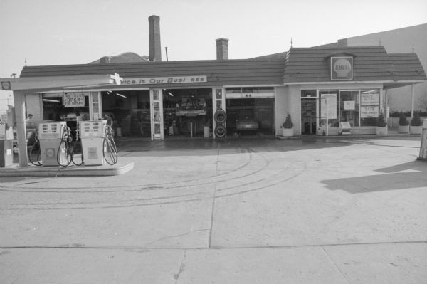 View across State Street towards the Shell Gas and Service Station. The four service doors are open, showing men working on cars. A sign above the gas pumps reads: "No Gas, Open for Repairs." Signs on the windows of the office on the right read: "'No' Gas;" "Radial's Belted's Conventional's;" and "'Low' Tire Prices."