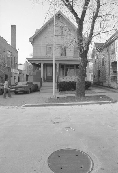 View across West Gilman Street towards a three-story house with a porch. A car is parked in the driveway on the left. The sign on the side of the house above the car reads: "No parking without consent from owner." A man is walking on the sidewalk. To the left of the house is an alley, and the rear of the 502 State Street building (Bigsby & Kruthers), and a sign above a door further down the alley reads: "Magic System, Insurance Agency Management." 