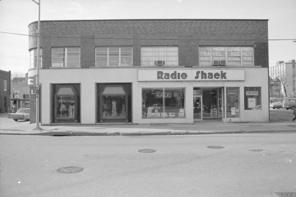 View across West Johnson Street, on the corner of State Street, towards a commercial building. On the right side, ground floor is Radio Shack. Above the Radio Shack is Hemispheric Travel Agency. On the left side ground floor are the show windows of Mulberry Bush Women's Clothing. Above the clothing store is office space for lease. The Radio Shack has signs in the window advertising a mid-winter sale. To the right of the building is a small parking lot.