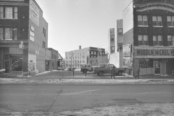 View across State Street towards an empty lot used for parking between Block Cleaners on the left, and the Wooden Nickel on the right. Three cars are parked in the lot. Billboards are on the side of the Wooden Nickel, and are advertising Pepsi and American Bank. There is a sign painted on the upper top side of the Block Cleaners building that is advertising the apartments for rent on the upper floors. Posters are on the side brick walls of both buildings. Pedestrians are walking on the sidewalk. In the background is of a house and apartment building on West Gilman Street (421 and 433 West Gilman Street).