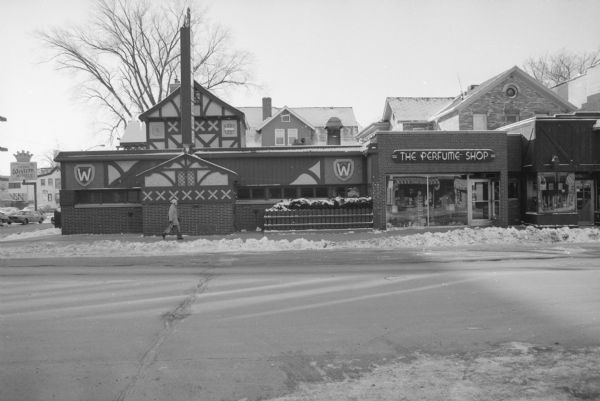 View across State Street, on the corner of North Frances Street, towards Brathaus (left) and The Perfume Shop (right). Snow is along the curb. Reflections of the buildings across the street are reflected in The Perfume Shop's windows. A pedestrian is walking by the Brathaus.