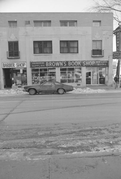 View across State Street, at the intersection of North Lake Street, towards a stone building with an Art Deco facade. The upper two floors are apartments. On the left side of the ground floor is a barber shop, and on the right is Brown's Book Shop. Signs for the bookstore are advertising school, office, and engineering supplies as well as new and used books. A man is walking on the sidewalk, and a car is parked along the snowy curb in front of the bookstore.