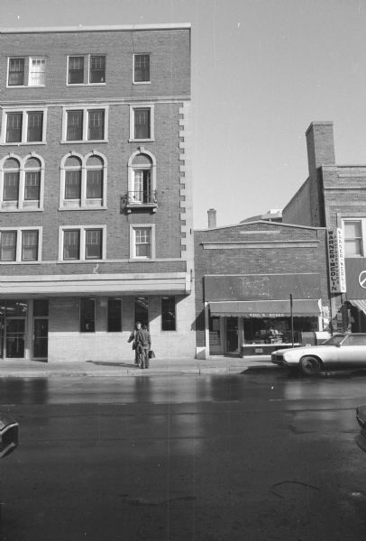 View across State Street towards the right side of the Rennebohm Drug Store building, and the building next door with Paul's Books on the ground floor. Two men are talking to each other on the sidewalk in front of the drugstore. Books are on display in Paul's Books show window. Two mailboxes are on the sidewalk in front of the book store, and cars are parked on both sides of the street. Along the right side of the bookstore is a sign for Warner Medlin Photographers.