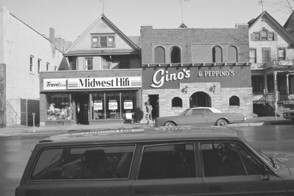 View across State Street towards two commercial buildings, with two houses behind them, and a house to the right of them. The commercial building on the left contains a travel center and Midwest Hifi, a radio and sound system store. The taller building on the right is Gino's & Peppino's restaurant, with a decorative brick facade and arched windows and doors. Two men are walking down the sidewalk past Gino's. Cars are parked on both sides of the street. 