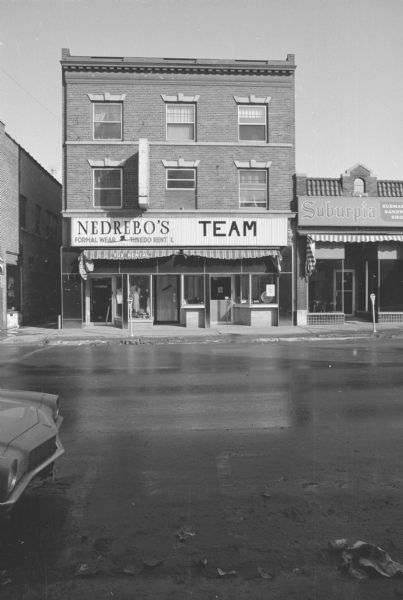 View across State Street towards the S.L. Parke Building. The ground floor contains two businesses: Nedrebo's Formal Wear and Tuxedo Rental on the left, with men's clothing on display in a show window. Team Electronics is on the right. On the far left across an alley is the Harry Wigonitz Building. To the right is the John Kelly Building.