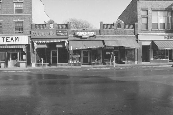 View across State Street towards the John Kelley Building. The facade is Spanish Colonial, and the building contains two businesses. Suburpia Submarine Sandwich Shoppe is located on the left side of the building. Meuer Art and Frame Company Co. is on the right side. On the left is the S.L. Parke Building, and on the right is the Hess and Schmitz Building.
