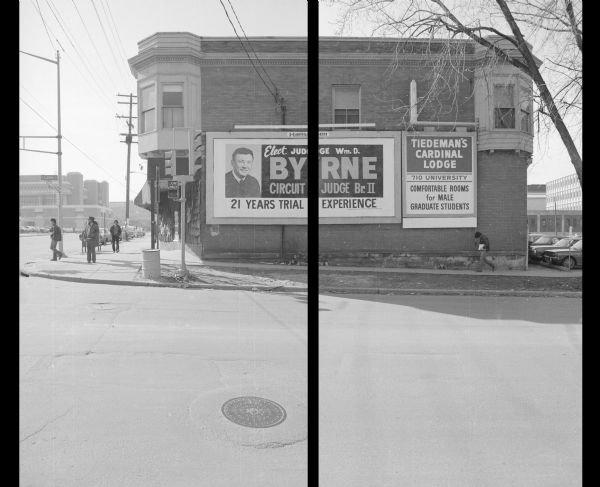 View across North Lake Street, on the corner of University Avenue, towards the Cardinal Building. Two billboards are on display on the side of the building. The one on the left reads: "Elect Judge Wm. D. Byrne Circuit Judge Br. II, 21 years trial experience." The one on the right advertises apartments in the upper floors of the building, and reads: "Tiedeman's Cardinal Lodge, 710 University, Comfortable Rooms for Male Graduate Students." On the left is a view down University Avenue. Pedestrians are walking down the sidewalk. Cars are parked in a parking lot on the right side of the building.