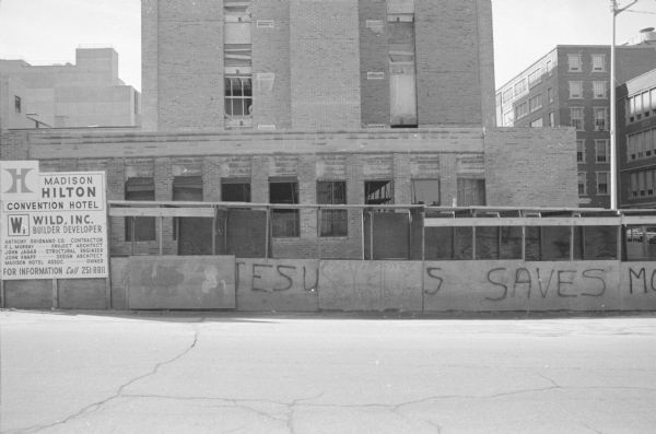 View across Wisconsin Avenue, on the corner of West Dayton, towards the construction of the Madison Hilton Convention Hotel. A sign on the left advertises the building and the construction company (Wild, Inc.). Graffiti on the scaffolding reads: "Jesus Saves M[?]."