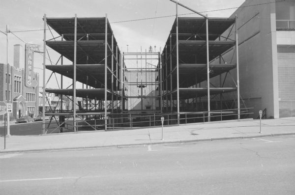 View across Wisconsin Avenue, on the corner of East Dayton Street, towards the construction of a parking ramp. Scaffolding is in place, and only a portion of the interior has been constructed. An electric sign on the corner of the ramp reads: "Pigeon Hole Parking."