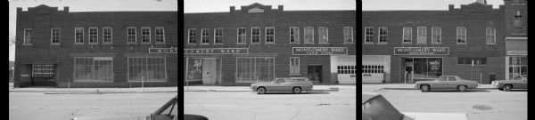View across North Henry Street towards the Montgomery Ward Farm Store. Several cars are parked along the curb in front of the long brick building. A sign in the window on the far right reads: "Mowers Repaired n Sharpened." The building, originally a car company and showroom, has two garage doors.