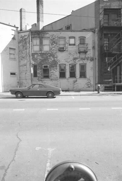 View across North Fairchild Street towards the back side of the Castle and Doyle Building, which houses the Castle and Doyle Fuel Company. Paint is peeling off of the facade, revealing the brick underneath. A car is parked at the curb in front of the building. The top of a parking meter is in the foreground.