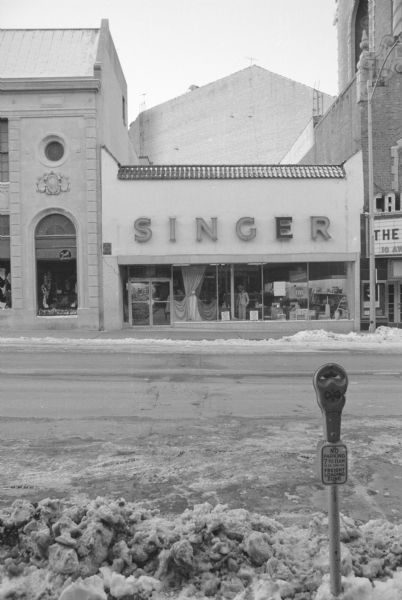 View across State Street towards Singer, a fabric and sewing store in a small building. Signs in the window are advertising a sale, and a mannequin is wearing a woman's pantsuit. To the left is the Yost-Kessinich Department Store, and to the right is the Capital Theater. There is a parking meter in the foreground.
