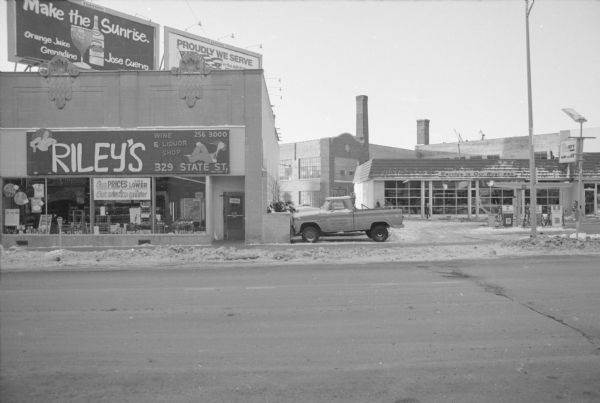 View across State Street, near the corner of West Gorham Street, towards Riley's Wine and Liquor Shop on the left, and a partial view of the Shell Gas Station on the right. Riley's is located in an Art Deco building. Behind the gas station is the Montgomery Ward Service Repair Department. A truck, belonging to the Shell Station, is parked in the service station lot next to Riley's. There are two billboards above Riley's roof, and the one on the left reads: "Make the Sunrise: Orange Juice, Grenadine, Jose Guervo," and the one on the right is only partially visible, and reads: "Proudly We Serve in the Air Force."