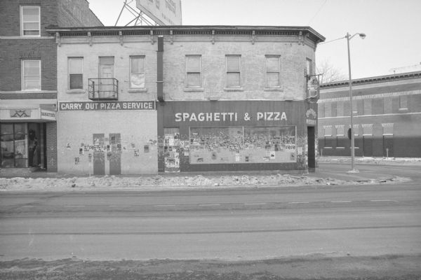 View across State Street, on the corner of North Broom Street, towards Gargano's Pizzaria. Electric or neon signs on the side of the building read: "Carry Out Pizza Service," and "Spaghetti & Pizza." Posters, flyers and the remains of removed posters cover the ground floor wall and window. A billboard advertising tequila is partially visible on top of the building. To the left is Martin's Midwest Western Wear. 