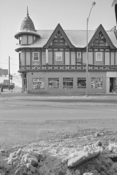 View across State Street, on the corner of West Gilman, towards the Stop 'N Go Grocery, a Tudor revival building with a turret on the corner. The far right and far left windows on the ground floor are covered in posters and flyers. Various goods are displayed in the middle three windows.