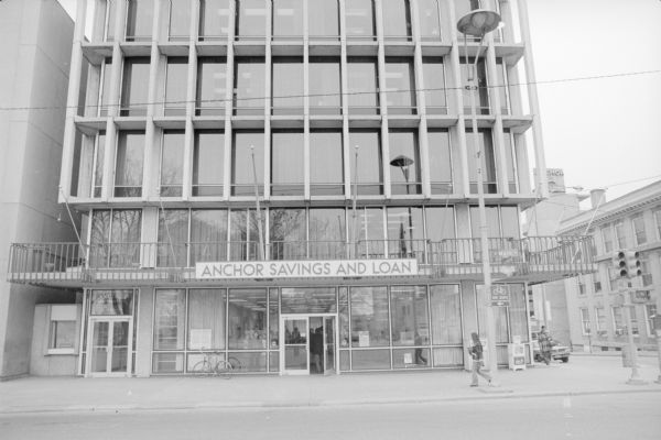 View across West Main Street, on the corner of South Hamilton Street, towards the Anchor Savings and Loan and office building. The largely glass building is reflecting the trees from Capitol Square across the street. Pedestrians are walking by on the street or into the building. One woman is standing inside the building on the ground floor looking out.