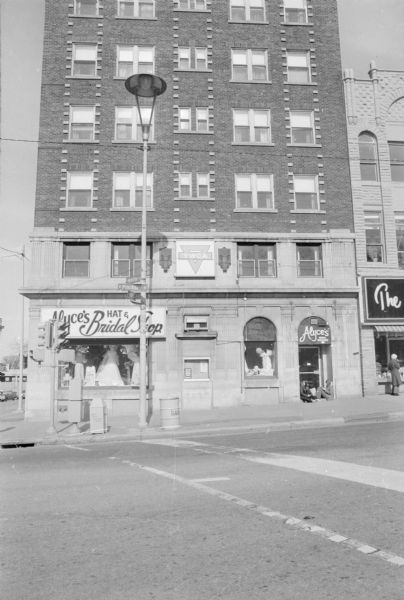 View across North Pinckney Street, where it intersects with East Mifflin and North Hamilton Streets, towards The YWCA, historically the Belmont Hotel, and the tallest building along Capitol Square. A sign with the YWCA's logo is on the second floor. The ground floor is Alyce's Hat & Bridal Shop. Mannequins wearing wedding dresses are in the window. Two young women are sitting in the doorway to Alyce's on the right. 