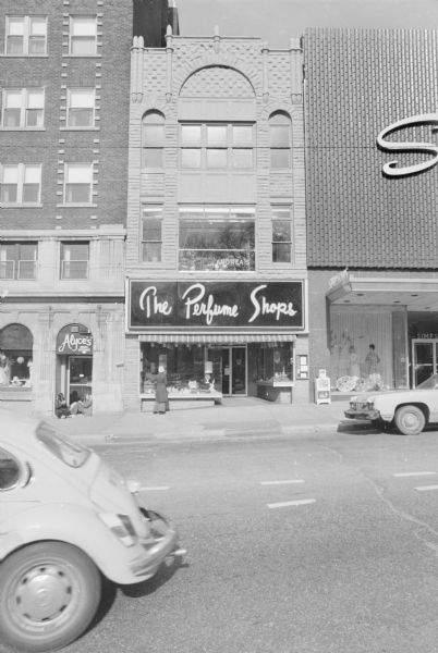 View across North Pinckney Street towards the Lydia Winterbotham Building, a Richardsonian Romanesque building. The ground floor is The Perfume shops, with display windows. A woman is standing outside the left display window and looking in. The second floor is Andrea's, a restaurant with numerous plants on display in the windows. To the left of the building, two women are sitting in the doorway to Alyce's Hat & Bridal Shop.