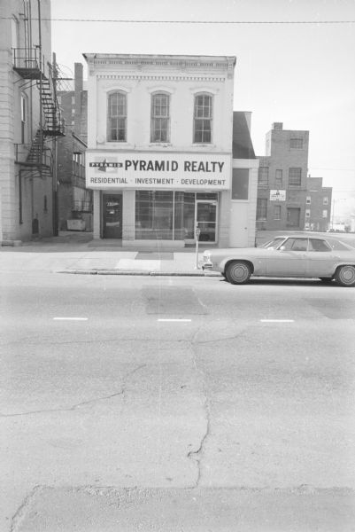 View across East Washington towards Pyramid Realty. A car is parked outside of the building. To the right is a small parking lot, and to the left is an ally between Pyramid Realty and the American Exchange Bank. A car is parked on the street in front of Pyramid Realty. The left most door (114 East Washington) has the words "Blied Printing Co." on it, as does a building in the background on the right.