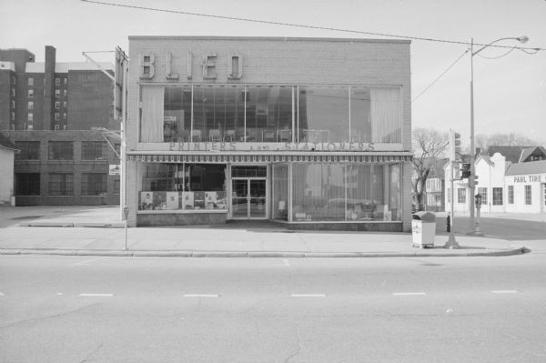 View across East Washington Avenue, on the corner of North Webster, towards Blied Printers and Stationers. Various items are on display in the ground floor windows. A line of chairs is visible through the second story window. To the left is a small parking lot. At the back of the parking lot is another building belonging to Blied Printers, and behind that building is the former Belmont Hotel (in 1973 the YWCA).