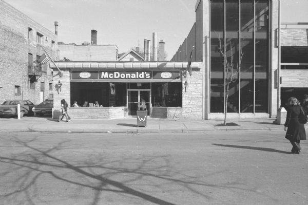 View across North Lake Street towards a McDonald's fast food restaurant. A woman is walking across the street on the right, and two young women are walking by the McDonald's on the sidewalk as a man is opening the door to go inside. Two flags are hanging from the building, one of them an American flag.