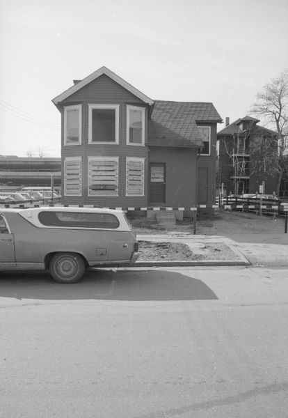 View across North Frances Street towards a house. The ground floor windows are boarded up, and the grounds are fenced off. On both sides of the house is an outdoor parking lot. An el Camino is parked on the street in front of the house.