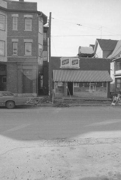 View across North Frances Street towards The Red Shed, a tavern in a small brick building. The windows are reflecting the buildings across the street. A covered wagon is sitting on the top of the building. The sidewalk in front of The Red Shed is torn and dug up, revealing the dirt underneath. To the left is the Olwell Building.