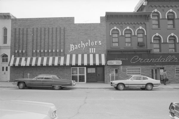 View across West Main Street towards two buildings. The one on the left is Bachelors III, a tavern. The Italianate style building on the right is the Mohr-Christoffer Block. The ground floor is Crandall's, another tavern. The upper floors are apartments. A sign over the left side door of Crandall's marks it as the entrance. Cars are parked on both sides of the street. To the left of Bachelors III is Smith and Lamb Block.
