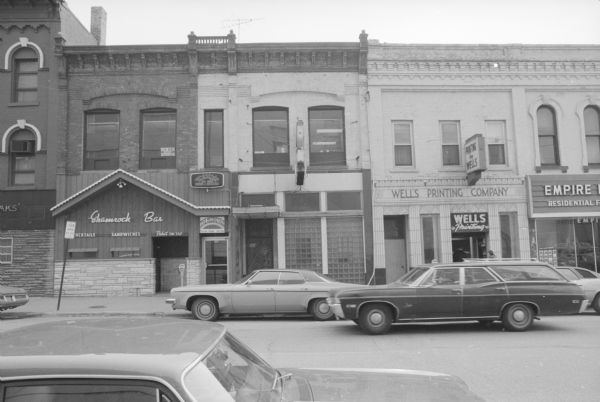 View across West Main Street towards two buildings. The Jacobs and Regan Blocks (two Italianate buildings built together) are on the left. The left side of this building is the Shamrock Bar, and the right side is the Paradise Lounge. A door between the two bars leads to The Rising Sun Sauna and Massage. Two signs mark the door, and one sign is electric. The upper floors are apartments, and a sign in the window above Shamrock Bar is advertising rooms for rent. On the left is the McGovern Block, an Italianate style building with an ornate art deco storefront. The ground floor is Wells Printing Company. To the right of these buildings is the Mohr-Christoffer Block. On the left, built to match the McGovern Block, is the Fitch Block.
