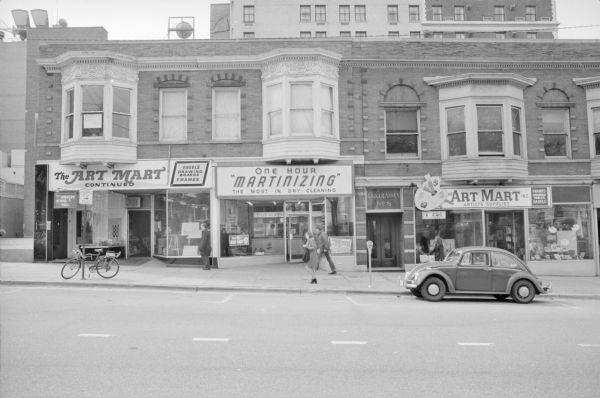 View across South Carroll Street towards two buildings. In full view on the left is the Badger Block, with decorative and ornate oriel windows. The ground floor contains two businesses: on the left is The Art Mart Continued, and on the right is One Hour Martinizing, a dry cleaner. Two signs, one above the leftmost door, and one in the leftmost oriel window above Art Mart Continued reads: "Innerlife School Yoga." The building on the right is part of the Badger Block Annex. It also has oriel windows, though with less decoration than the Badger Block. The business on the ground floor is The Art Mart, Inc. Pedestrians are walking on the sidewalk, and a bicycle is leaning against a street sign. A Volkswagen Beetle is parked along the curb on the right.