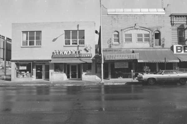 View across State Street towards two buildings. On the far left is the back of a large billboard next to the side of the brick building which has two businesses on the ground floor, Electric Shaver Service and Schwartz Jewelers. Schwartz Jewelers has a large electric or neon sign, including the shape of a diamond, above the door. Potted plants are inside the window on the second floor. The building on the right is the H. Halperin Building, with two businesses on the ground floor. On the right is The Consignment Shop. The awning above the store reads "Undertaking" which was the name of the health food store that had been in business at that location the year before. The business on the right is The Medium, with faded lettering on the awning which reads: "The Black Market." The proprietor of The Medium specializes in tarot, palmistry, and horoscopes.