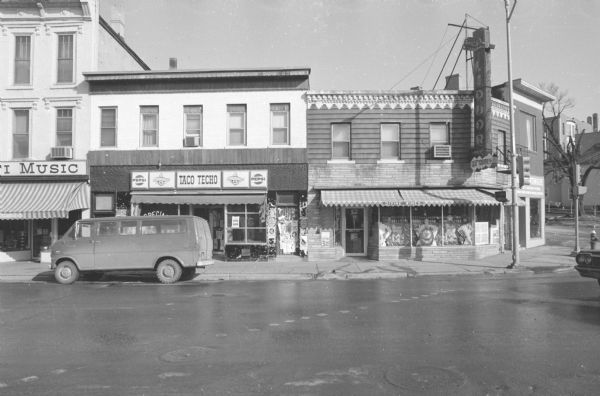 View across State Street, at the intersection with West Gorham Street, towards two buildings. The building on the left is the M.B. French Building, with Taco Techo on the ground floor, with osters and flyers covering a door on the left side of the building,. There is a van parked along the curb in front. The building on the right is Badger Liquor, with a large neon sign on the corner, and liquor bottles in a decorative display in the windows.