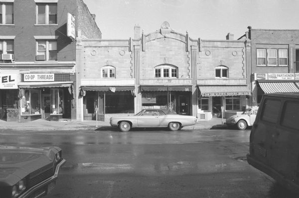 View across State Street towards the Bertrand Building, a Neoclassical style two-story building. The ground floor consists of three businesses. The far left appears to be an empty storefront (it had been Compared to What? a shoe boutique). In the middle is Shakti Bookshop. The far right is Klein's Bakery. Cars are parked on both sides of the street.