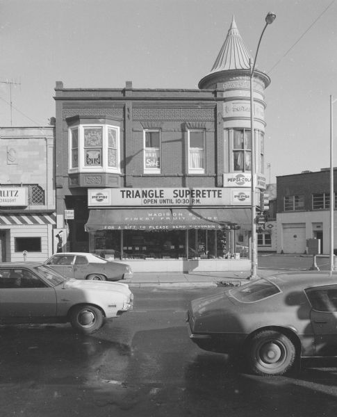 View across State Street, at the intersection with North Henry Street, towards the Queen Anne-style Matthew Gay Building, a two-story building with a turret above the corner entrance. The ground floor is Triangle Superette, with advertisements for Pepsi on either side of the store name. Words on the awning read: "Madison's finest fruit store," and "For a gift to please send Wisconsin cheese." Fruits, cheeses, and other foods are on display in the ground floor window. The second floor is Yellow Tower, a clothing store. The oriel window on the second floor on the far left is decorated and painted to advertise Yellow Tower. A sign below the window is marking the door that leads to the second floor. Cars are driving by or are parked along the curb in front of the building.
