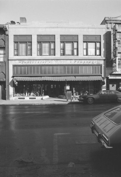 View across State Street towards a two-story stone commercial building. The ground floor is Rentschler Floral Company. Flowers and potted plants are on display in the windows on either side of the door. A sign in the left ground floor window is advertising St. Patrick's Day cards. The cross at the top of Holy Redeemer Catholic Church (behind the store and across West Johnson Street) can be seen above the building. Cars are parked on both sides of the street. To the left is the Conklin Block. To the right is Nick's restaurant, with an electric sign advertising the restaurant.
