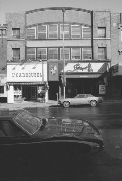 View across State Street towards the three-story Goodman Building. The upper two floors are apartments. The ground floor contains two businesses. On the left is Le Carrousel European Fashion. On the right is The Clothes post, with Charles Speth Associates on the upper floors. The Clothes post is moving, and there is a sign in the window advertising the store space for lease. The crest or logo for Charles Speth Associates is above the door between the two stores. Cars are parked on both sides of the street. To the left is the Fred Rentschler Building, and to the right is the Anna Weber Building.