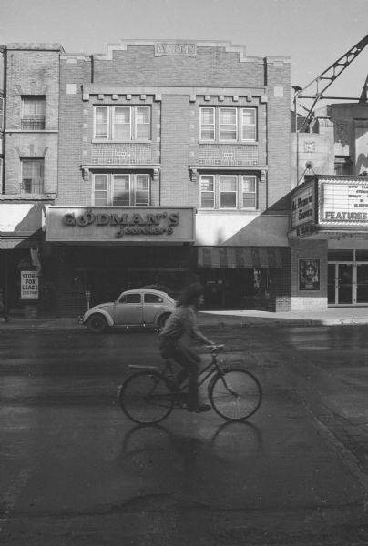 View across State Street towards the Anna Weber Building. The upper two floors are apartments. The ground floor is split into two businesses: on the left is Goodman's Jewelers, on the right is Midwest Camera. A car is parked in front of Goodman's Jewelers, and a man is riding a bicycle down the street in the foreground. To the left of the building is the Goodman Building, and to the right is the Orpheum Theater.