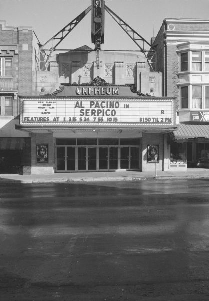 View across State Street towards the Orpheum Theater, an art deco building with a large electric sign and a marquee. The marquee reads: "Al Pacino in Serpico." Two movie posters for Serpico frame the doorway under the marquee.