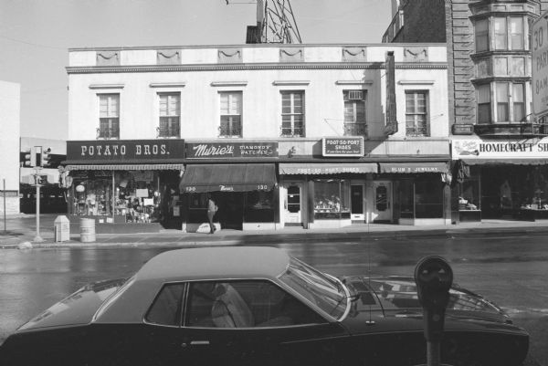 View across State Street, at the intersection with West Dayton, towards the neoclassical two-story Stanley Hanks Building. Four businesses make up the ground floor. From left to right they are: Potato Bros., a general store; Murie's, diamonds and watches; Foot-So-Port Shoes; and Blum's Jewelery. All of the businesses have awnings, and their goods on display in the show windows. Blum's Jewelry has a large electric sign above the entrance between two windows. In the foreground a car is parked at a parking meter.
