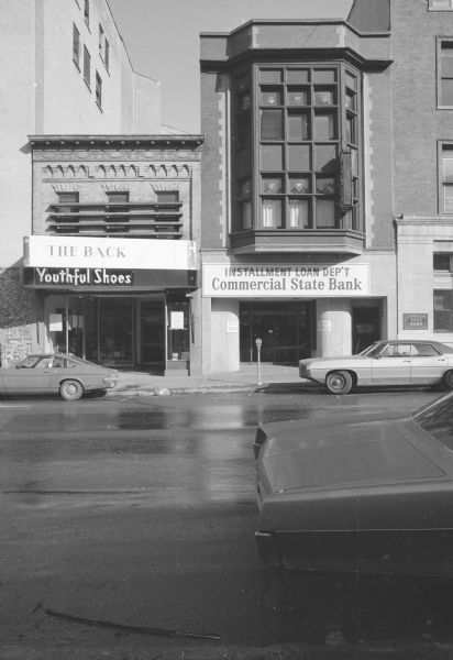 View across State Street towards two buildings. The two-story building on the left is the Mautz Brothers Block, and on the ground floor is Youthful Shoes, with show windows flanking the door. The three-story Queen Anne style building on the right is the Lamb Building. The ground floor is Installment Loan Department of the Commercial State Bank. The oriel windows of the second and third floor include leaded and stained glass. An electric sign attached to the second floor reads: "Music." Cars are parked on both sides of the street.