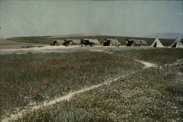 Red poppies and other wildflowers are blooming in profusion near a camp of seven tents. Towels or laundry are hanging from clotheslines, and there is an automobile parked in front the tents. Two men are sitting on folding chairs on opposite sides of a small table. There are hills in the background.
