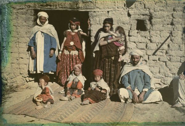 A group of two men, two women and four children in traditional dress are posing in front of the door of a brick dwelling. Three of the children are sitting on a rug on the ground, and one of the women is holding the other child in her arms.