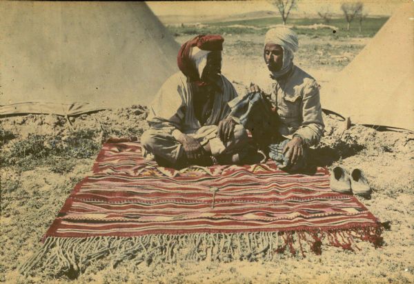 Two men are smoking cigarettes outdoors. They are wearing turbans and traditional dress, and are sitting on a colorful rug with long fringe.  A pair of shoes is on the left at the edge of the rug, and what may be a pocket watch is resting in the center of the rug. There are two tents behind the men, and trees and hills in the background.