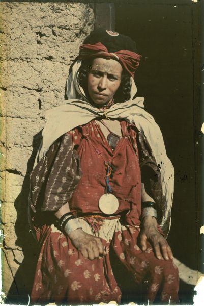 A woman is posing sitting in the doorway of a brick building. She has facial tattoos and is wearing a head scarf over her braided hair.